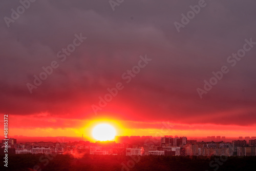 Fototapeta Naklejka Na Ścianę i Meble -  Hot red sun behind the silhouettes of city buildings. Summer sunset in the sky haze with night houses