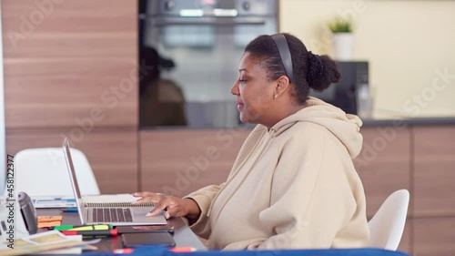 Tracking shot of plus size Black woman sitting down at desk and having video call on laptop from home. Female finishing talk, closing pc, looking at camera and smiling. Remote work and self-isolation