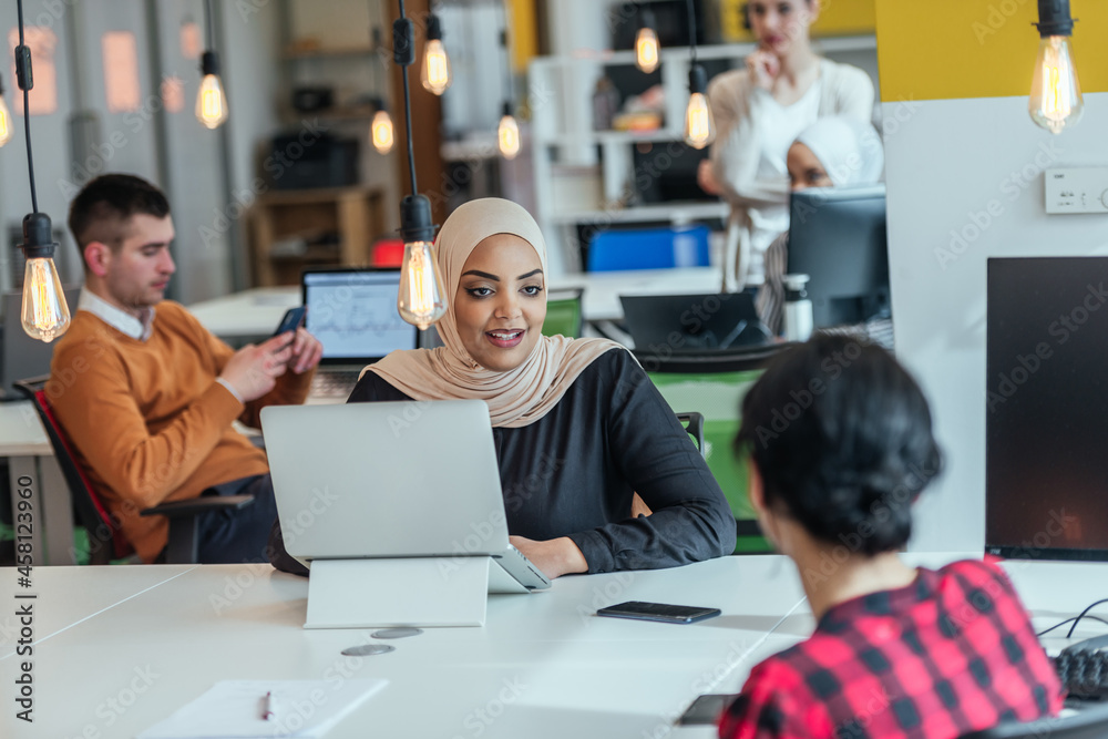 Group of coworkers, teammates helping their new African American, black, muslim colleague wearing hijab to integrate into the modern company. Multiethnic colleagues in a modern startup company. Group of coworkers, teammates helping their new African American, black, muslim colleague wearing hijab to integrate into the modern company. Multiethnic colleagues in a modern startup company.
