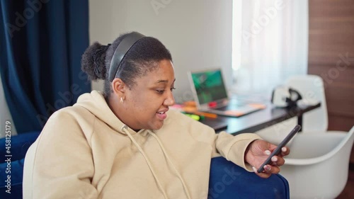 Portrait of chubby Black woman in beige hoodie typing sms on cellphone, looking at camera and smiling. Young female text messaging on phone and posing sitting on sofa while working from home photo