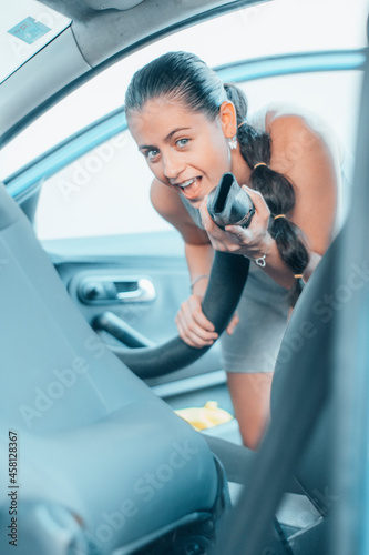 Chica joven feliz bromeando con la aspiradora mientras limpia su vehículo en el túnel de lavado de coches © Migue Suarez