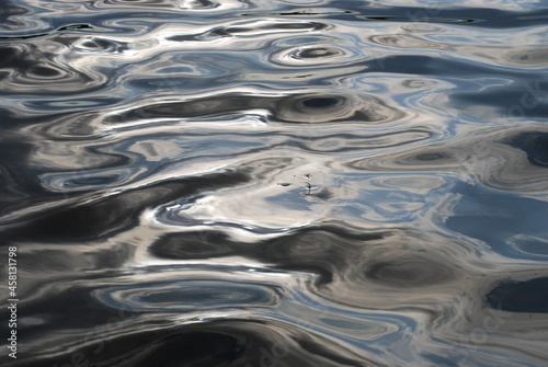 Small waves on the water. On the surface of the water there are smooth waves of different shapes. Light blue skies and white clouds are reflected from them, creating an abstract blurry picture.