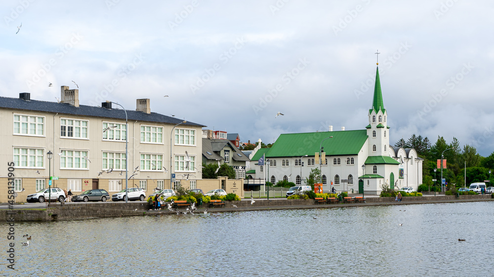 Obraz premium Beautiful view of the Iceland Church with white walls and green roof in front of the lake on a cloudy sky in Reykjavik, Iceland. Architecture, landmark, worship service, religion, faith concept