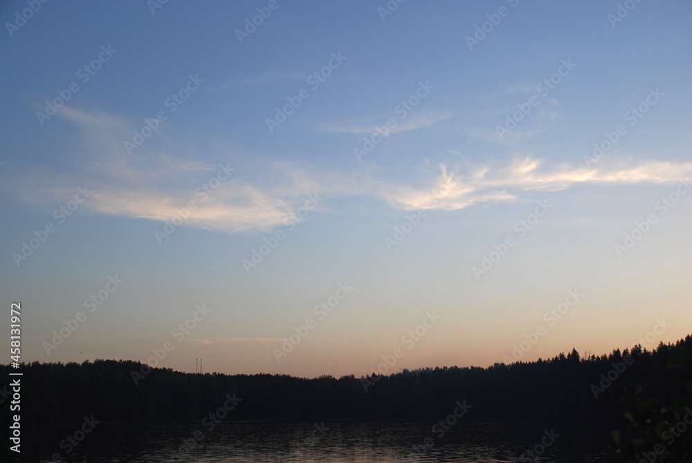 A white, weightless cloud spread across the sky. A semi-transparent white airy barely perceptible cloud stretched out against the background of a light blue sky over a wide strip of dark forest.