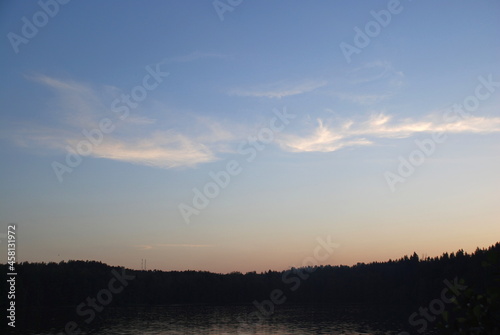A white  weightless cloud spread across the sky. A semi-transparent white airy barely perceptible cloud stretched out against the background of a light blue sky over a wide strip of dark forest.
