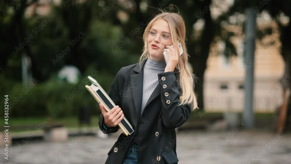 Young girl in glasses with books walking near the university building. Portrait of attractive student having phone talk. Happy and smiling and walking forward. High quality 4k footage Young girl in glasses with books walking near the university building. Portrait of attractive student having phone talk. Happy and smiling and walking forward. High quality 4k footage