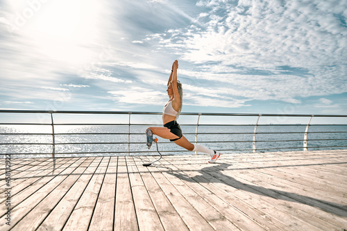 Life balance. Calm and sporty disabled athlete woman in sportswear with prosthetic leg standing in yoga pose on the bridge in front of the sea.