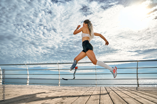 Obraz na płótnie Full length portrait of a confident young disabled sportswoman running fast on the bridge near the sea