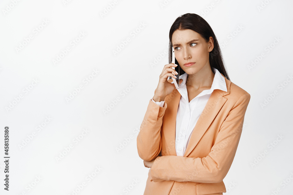 Serious corporate woman talking on mobile phone, frowning and listening with concerned face, standing in suit over white background Serious corporate woman talking on mobile phone, frowning and listening with concerned face, standing in suit over white background