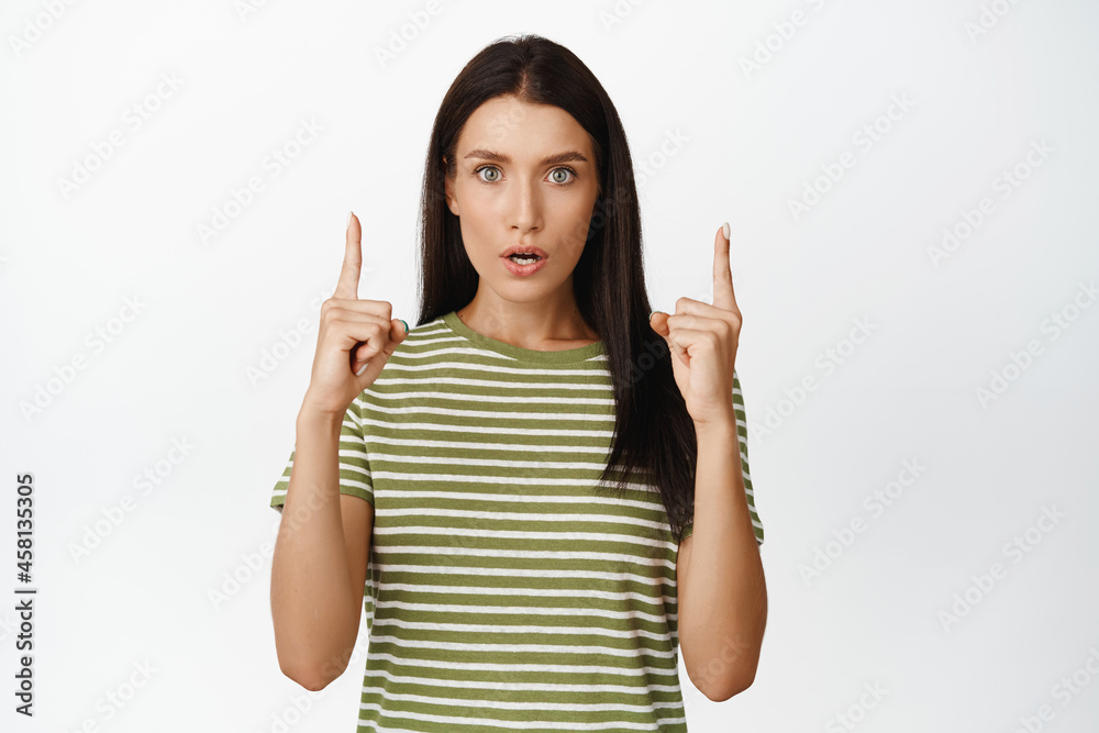 Image of surprised adult woman pointing up, looking in awe at camera, standing in t-shirt over white background Image of surprised adult woman pointing up, looking in awe at camera, standing in t-shirt over white background