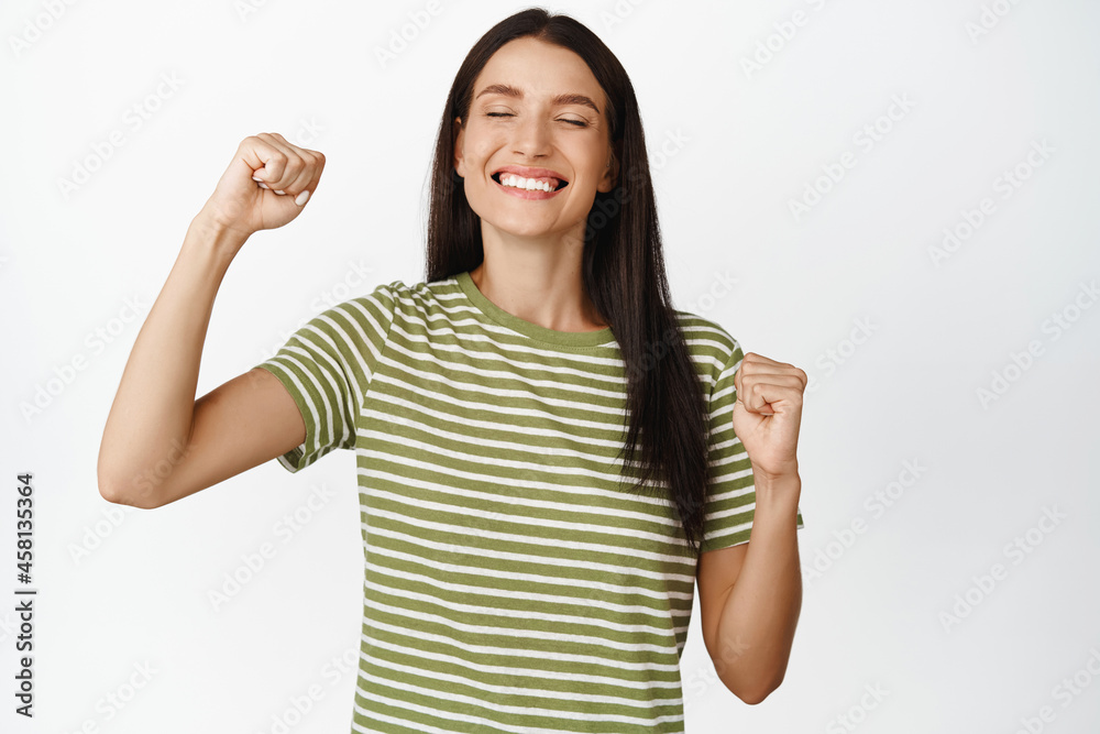 Excited smiling woman close eyes, standing with pleased face, makes fist pumps, winning, achieve goal and celebrating, standing over white background Excited smiling woman close eyes, standing with pleased face, makes fist pumps, winning, achieve goal and celebrating, standing over white background