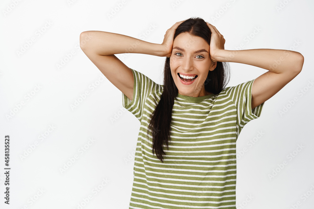 Happy brunette woman looking excited, smiling at camera, holding hands on hair, standing in tshirt over white background Happy brunette woman looking excited, smiling at camera, holding hands on hair, standing in tshirt over white background
