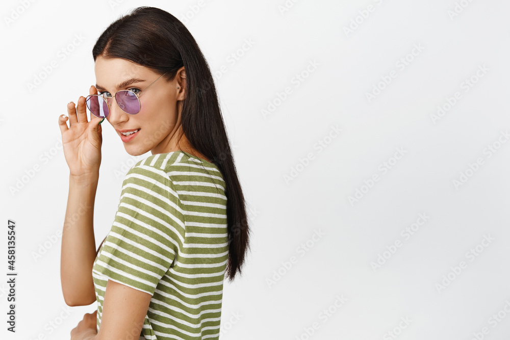 Sassy brunette girl in sunglasses, gazing at camera flirty, standing in t-shirt over white background