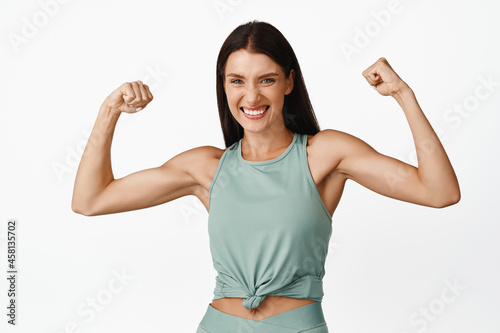 Strong beautiful woman flexing biceps, showing muscles on arms and smiling happy, bragging with fit and healthy body, concept of workout and exercises