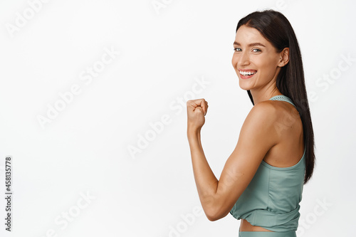 Strong young fitness woman showing muscles on arm, flexing biceps and smiling pleased, sharing workout result, standing over white background