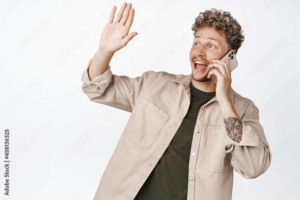 Friendly handsome man talking mobile phone and saying hello to friend, waving hand to greet person aside, standing in casual clothes over white background Friendly handsome man talking mobile phone and saying hello to friend, waving hand to greet person aside, standing in casual clothes over white background