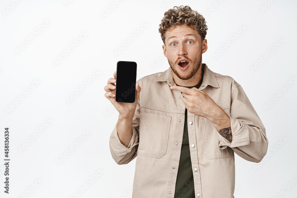 Excited blond curly guy pointing finger at mobile phone screen, looking surprised, showing awesome news online, standing over white background Excited blond curly guy pointing finger at mobile phone screen, looking surprised, showing awesome news online, standing over white background