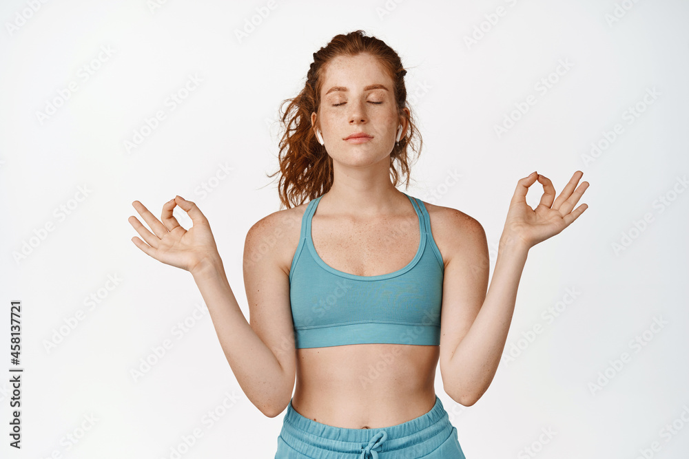 Calm and relaxed redhead girl meditating, control breathing, close eyes and practice yoga, standing in activewear against white background Calm and relaxed redhead girl meditating, control breathing, close eyes and practice yoga, standing in activewear against white background