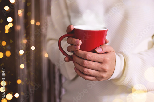 Merry Christmas. Woman in white sweater hands holding christmas hot drink or cofee in red cup. Shimmer background with snow and light bokeh. Greating card or banner