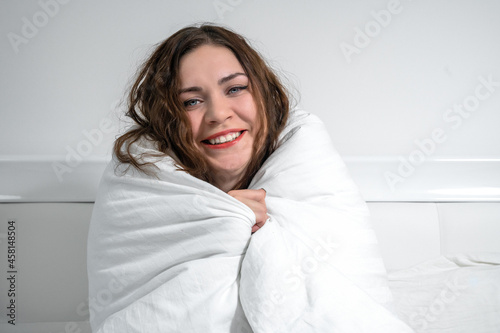 Attractive young brunette woman has wrapped herself in white duvet to keep warm and is laughing, while sitting on the bed. She feels the approach of winter. Lack of heating at home in cold season.
