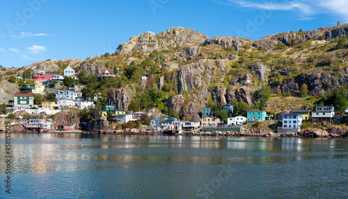 The hillside of St. John's Harbour, on a sunny day, under blue sky and white clouds. The colorful wooden houses are scattered along the hillside with the blue ocean in the foreground. 