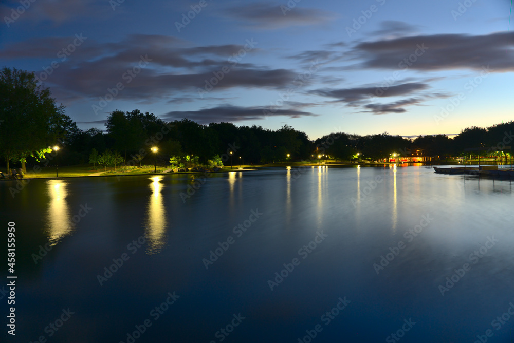 A long exposure of Beaver Lake, Mount Royal, Montreal, QC, Canada at twilight blue hour on a cloudy summer day. The lights and trees reflect on the lake.