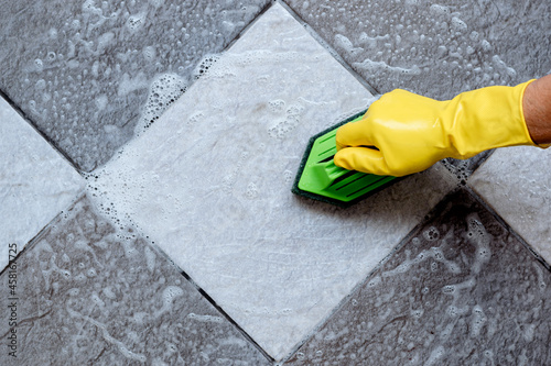 Top view of human hands wearing yellow rubber gloves are using a green color plastic floor scrubber to scrub the tile floor with a floor cleaner.
