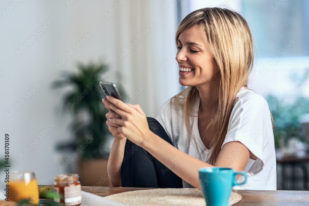 Smiling young woman enjoying breakfast while using her mobile phone in the living room at home. Smiling young woman enjoying breakfast while using her mobile phone in the living room at home.