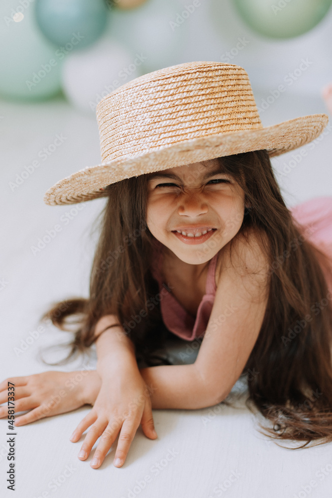 Beautiful smiling little dark-skinned girl with brown eyes and dark hair in a straw hat and a dusty pink jumpsuit is lying on the floor against the background of colorful balloons.