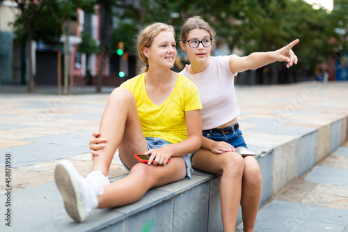 Youth positive girls sitting in city park together. One girl pointing index finger and looking in direction of it.