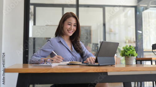 Attractive businesswoman using computer tablet and making notes on notebook.