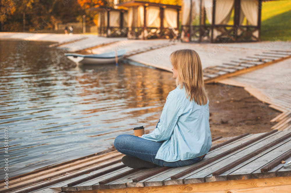 Autumn relax. A blonde woman sits on the shore of an autumn pond with a glass of hot coffee or tea in her hands. View from the back. A place for text. Autumn relax. A blonde woman sits on the shore of an autumn pond with a glass of hot coffee or tea in her hands. View from the back. A place for text.