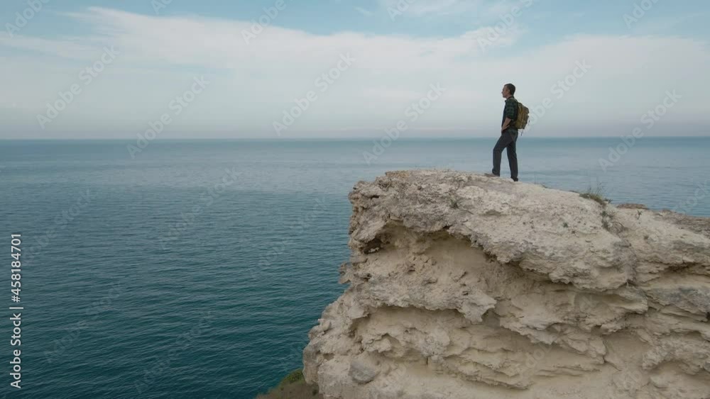 one person male with backpack standing on edge of rocky mountain on blue sky and sea horizon background shot by flying drone aerial view camera movement one person male with backpack standing on edge of rocky mountain on blue sky and sea horizon background shot by flying drone aerial view camera movement
