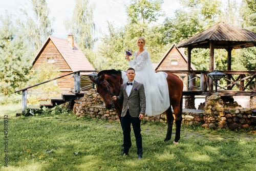 Wedding. Portrait of happy young married couple, beautiful bride in white dress with bouquet sits on horse, groom standing nearby, smiling newlyweds posing, looking at camera. Selective focus on man © Sergio