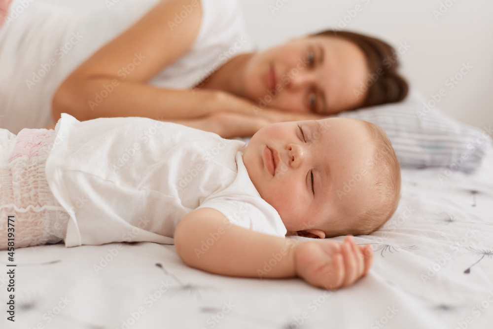 Indoor shot of attractive dark haired female wearing white t shirt lying on bed with her infant baby, having rest together, mommy looking at her cute daughter. Indoor shot of attractive dark haired female wearing white t shirt lying on bed with her infant baby, having rest together, mommy looking at her cute daughter.