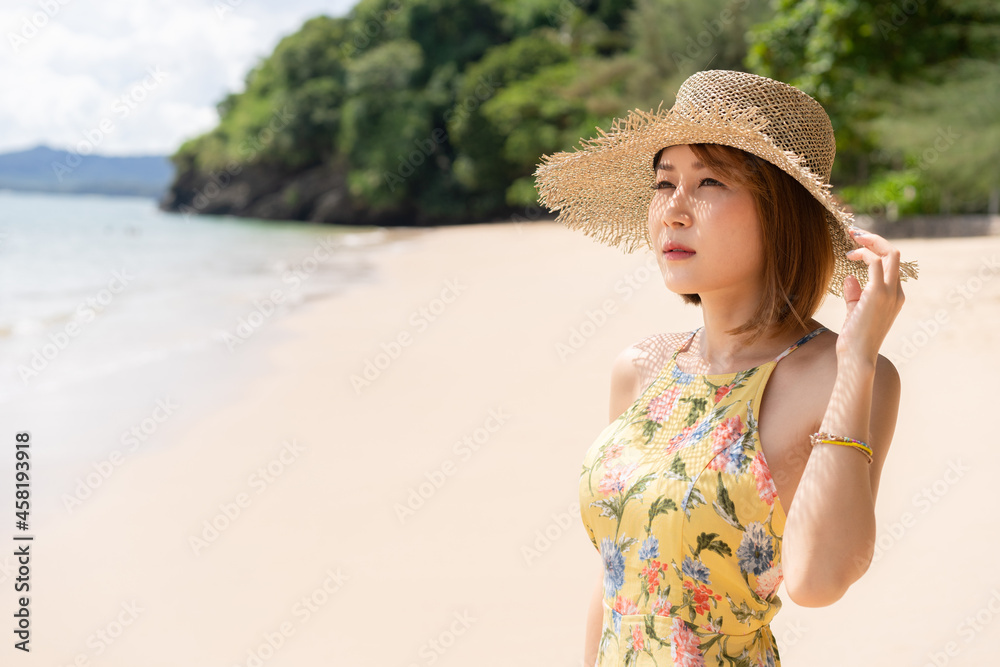 Young short-haired Asian woman in a yellow floral dress and straw hat standing on beach with one hand touching hat admired beautiful seascapes with a blurry mountain blue sky background on a sunny day
