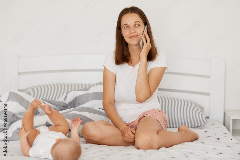 Dreamy smiling woman wearing white t shirt and shorts talking via cell phone and looking away, sitting on bed near her little daughter, family posing together at home. Dreamy smiling woman wearing white t shirt and shorts talking via cell phone and looking away, sitting on bed near her little daughter, family posing together at home.