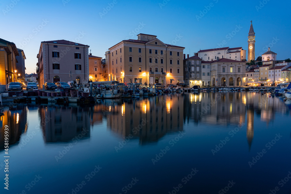 Piran harbour in the evening at blue hur with fishing boats and illuminated buildings saint george church tower