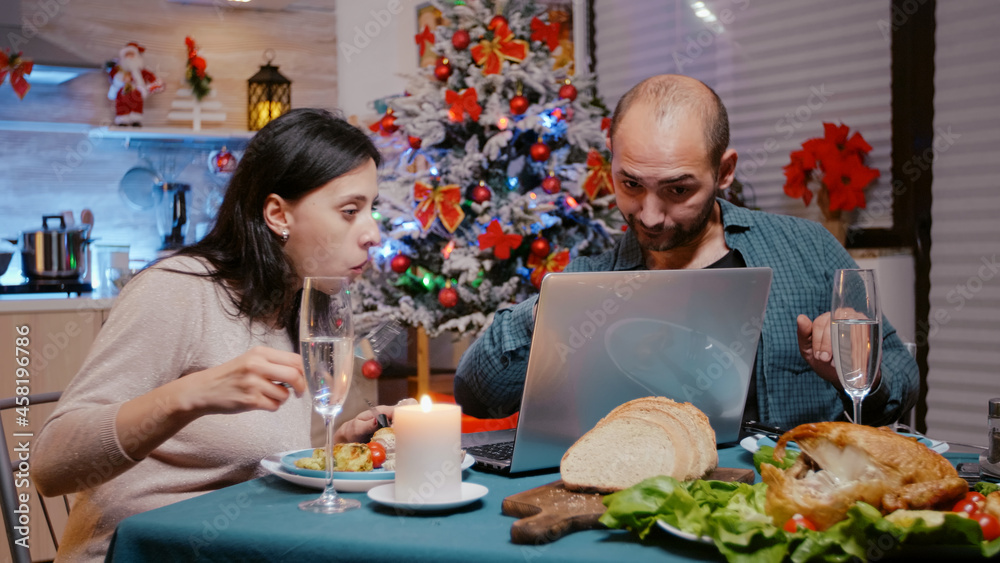 Festive couple looking at laptop and enjoying christmas dinner with technology. People eating chicken and drinking champagne while using device. Man and woman celebrating holiday.