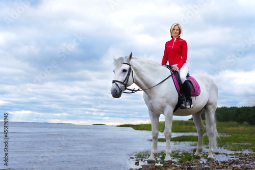 Portrait of a beautiful girl, young woman rider, equestrian on white horse in polo shirt, riding outdoors near lake or beach gulf. Horseback riding sport, natural background.
