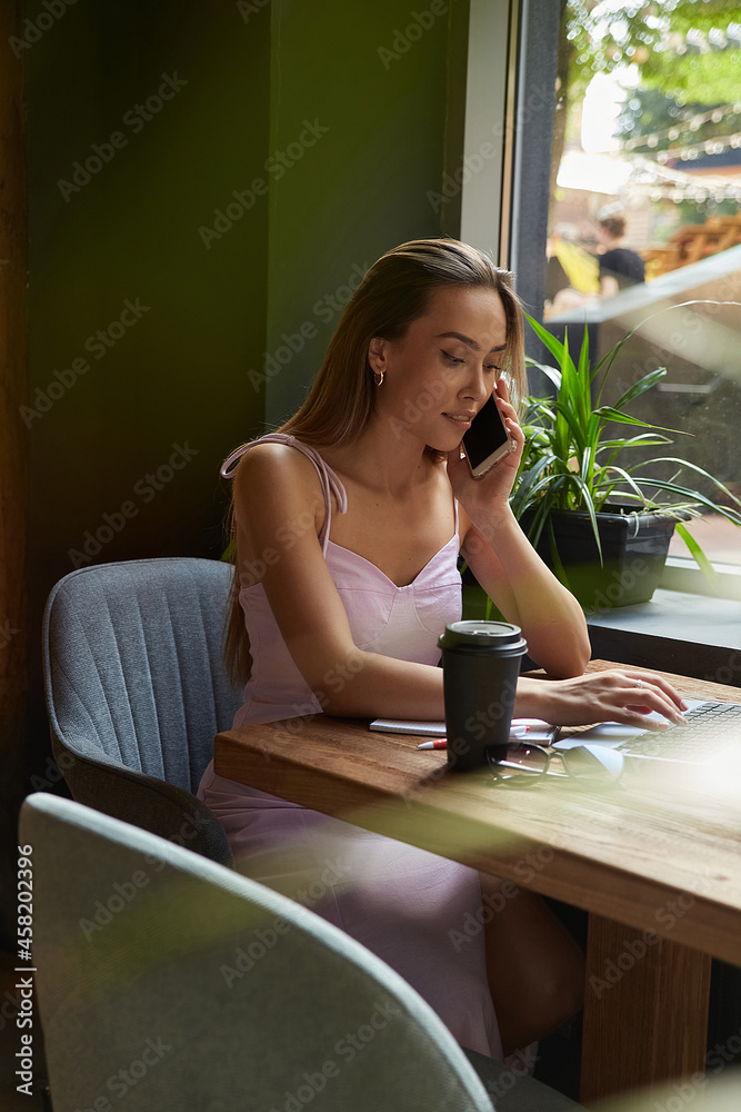 beautiful young asian woman sitting at coffee shop table surrounded by plants, working on laptop. beautiful lady calling by smartphone. modern online education, distant work, remote job, freelance beautiful young asian woman sitting at coffee shop table surrounded by plants, working on laptop. beautiful lady calling by smartphone. modern online education, distant work, remote job, freelance