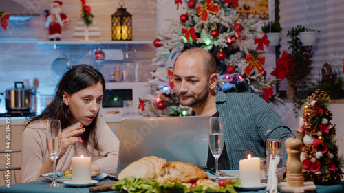 Couple doing online shopping on laptop with credit card for christmas presents while eating festive dinner. Man and woman buying gifts on internet for holiday celebration  enjoying meal.