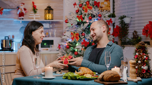 Couple celebrating christmas eve with presents while enjoying festive dinner. Man giving gift to cheerful woman and eating traditional food for holiday celebration. Seasonal festivity