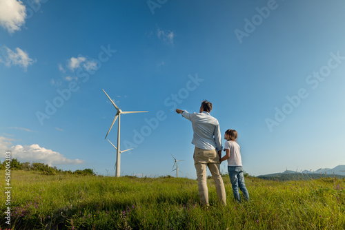 Cinematic shot of carefree young father engineer keeping his daughter for hand and showing windmill field. Concept of renewable energy, love for nature, family, electricity, green, future. © Kitreel