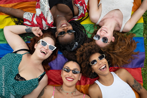 Cinematic shot of girl friends of different ethnicities with sunglasses while lying on LGBT rainbow flag in park. Concept of friendship, homophobia, diversity, equality, freedom, liberty, multiethnic.