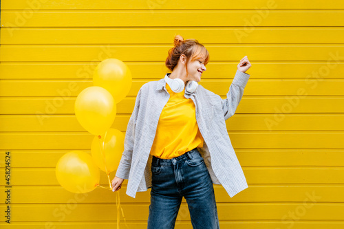 Festive occasion, holiday celebration concept. Adult hipster girl enjoy life, dancing with helium yellow balloons on yellow background in the city street outdoor.