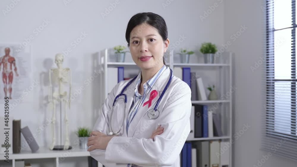 portrait female medical worker standing with her arms crossed is smiling at camera with a pink ribbon for breast cancer awareness at a bright clinic. portrait female medical worker standing with her arms crossed is smiling at camera with a pink ribbon for breast cancer awareness at a bright clinic.