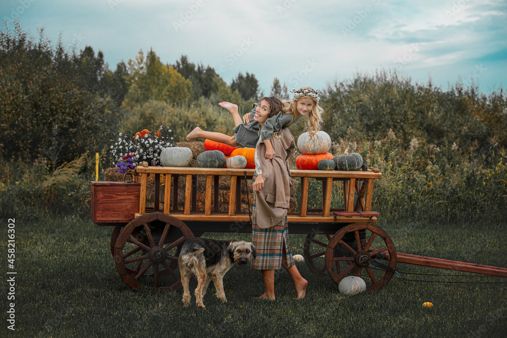 Obraz premium Beautiful happy family, mother, daughter and pet dog together, on a wooden cart with colorful pumpkins