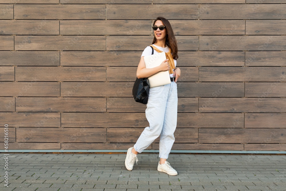 Full length photo shot of young charming beautifyl brunet woman going in the street near brown wall looking to side in white t-shirt and light blue jeans white sneakers with black female bag holding Full length photo shot of young charming beautifyl brunet woman going in the street near brown wall looking to side in white t-shirt and light blue jeans white sneakers with black female bag holding