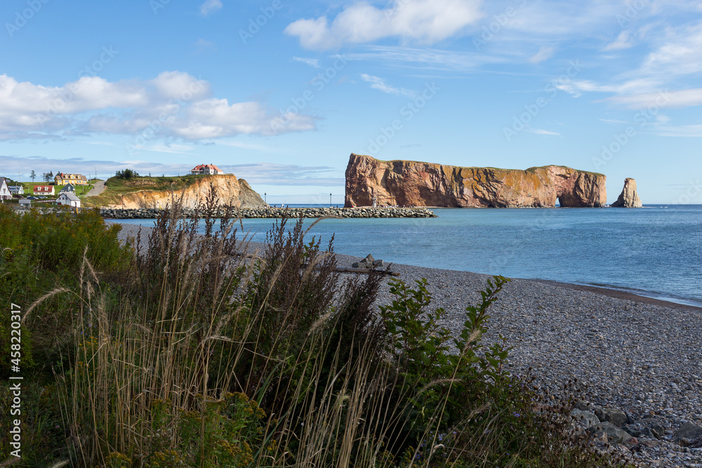 Grasses on beach with the famous limestone Percé Rock and a scattering of houses in the village seen during a beautiful afternoon, Quebec, Canada